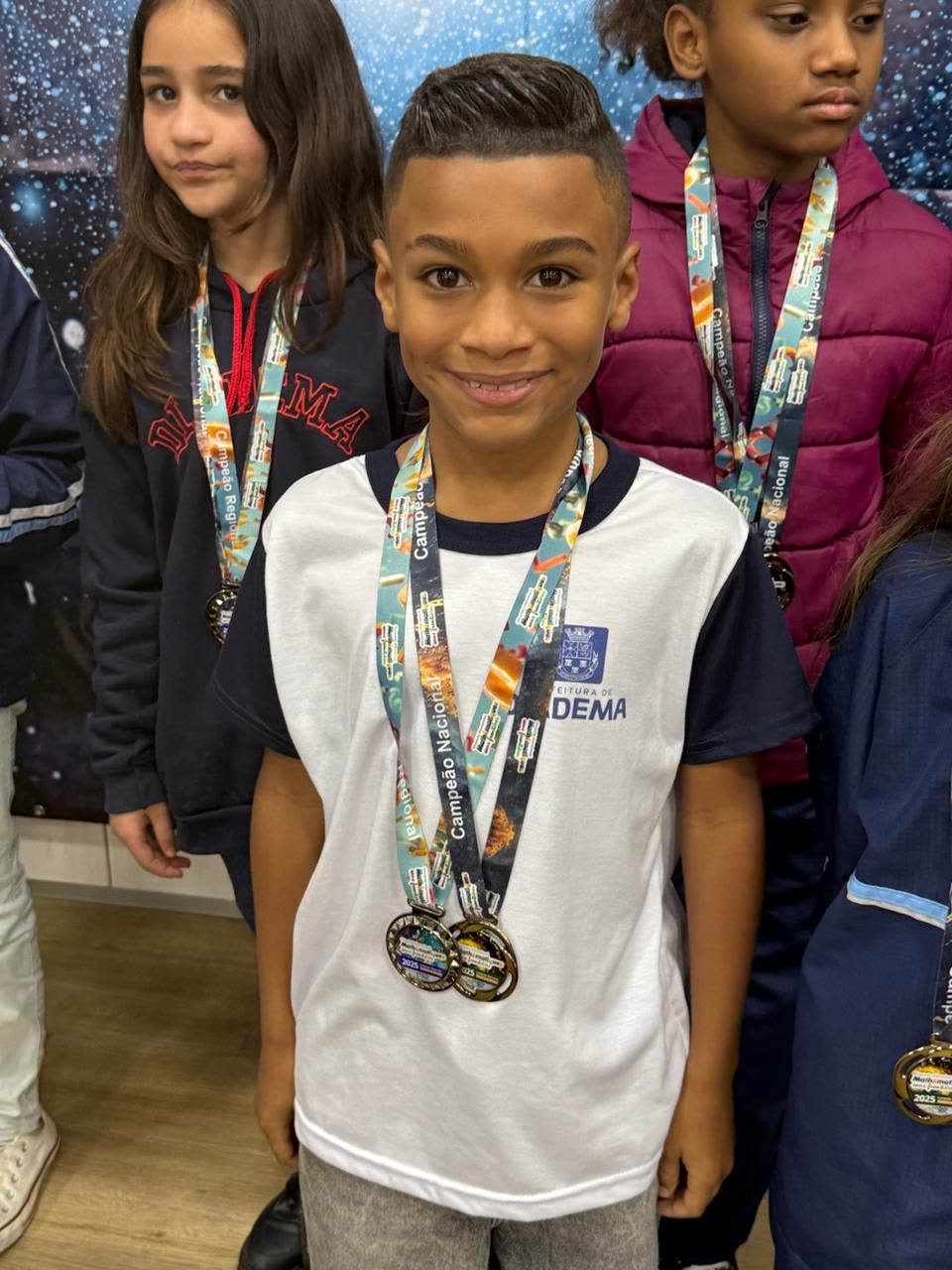 Estudante sorrindo com duas medalhas no peito, colegas ao fundo